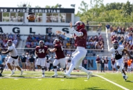 A football player in a red jersey and helmet throws a football across a field of other players as a crowd looks on