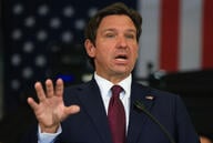 A photo of Florida Governor Ron DeSantis, a white man with brown hair, wearing a dark suit with a maroon tie and gesturing with his right hand.