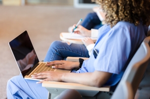 An unidentified female student uses a laptop to take notes in class. She is wearing scrubs and is sitting on the front row.
