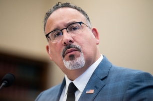 Education Secretary Miguel Cardona, a middle-aged Hispanic man with glasses and a salt-and-pepper goatee, wears a blue suit at a House budget hearing.