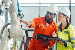 An instructor in a hard hat instructs a student in a hard hat, pointing to a machine.