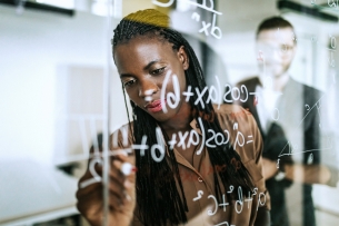 Minority woman writes equation on transparent board while instructor looks on behind her