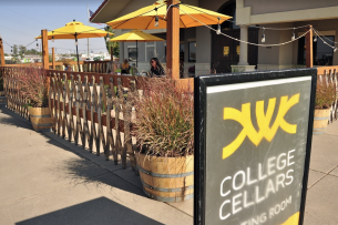 Two people sit in the background of a winery patio under a yellow umbrella. Sign in front of frame says College Cellars.