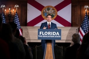 Governor Ron DeSantis speaks at a podium in front of the Florida state flag. A blue sign with white lettering on the podium reads: “Florida: The Education State.”