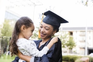 A woman in a graduation gap and gown holds a little girl.