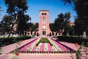 A University of Southern California campus photo, depicting a long walkway toward the entrance of a USC building. A garden with pink and white flowers and well-manicured shrubbery bisects the brick walkway.