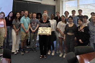 A group of college students surrounding their instructor, in front of a screen with the name of the course on it.