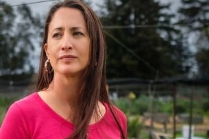 Elizabeth Hoover, a light-skinned woman with long dark hair, is wearing a pink shirt and standing in a garden.