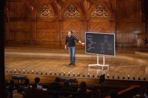 A man in a black sweater in a wooden lecture hall gestures to a blackboard with a flow chart on it.