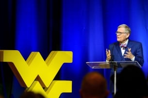 A man in glasses and a bow tie speaks at a podium in front of a blue curtain with a yellow West Virginia University emblem on the left side of the photo.