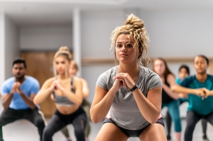 Photo of young people in a fitness class, squatting. Focus is on a young woman in front with her hair in a bun.