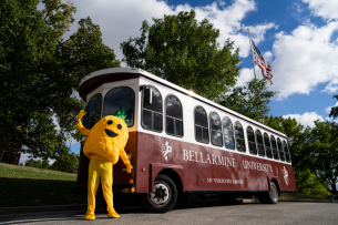 A pineapple mascot stands in front of a trolley with Bellarmine University printed on the side.