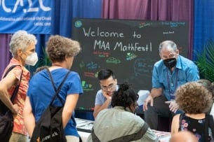 Six people, including at least two wearing masks, gather around a table with "Welcome to MAA Mathfest!" written on a chalkboard behind them.