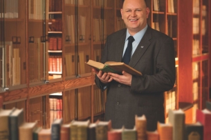 Alan Bearman, a bald man with light skin wearing a business suit, smiles holding a book in a library.