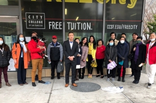 City College of San Francisco board president Alan Wong stands in a row with a group of representatives of organizations supporting Cantonese programming in front of a college building on the Chinatown campus. Many of the people are Asian American, and some are wearing blue paper masks.