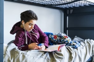 A student lies on their dorm bunk with a book open, on their phone