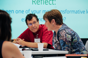 Attendees of a conference, two middle-aged light-skinned people, talk at a table.