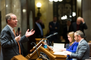 A man in a suit and tie speaks at a podium to a room of legislators