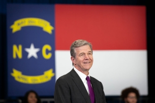 A light-skinned man in a suit and tie with gray hair stands in front of the North Carolina state flag