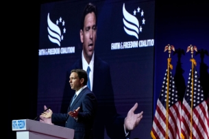 Florida governor Ron DeSantis stands at a podium in front of two American flags. His image is displayed on a big screen behind him.