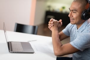 Man with headphones sits a desk with his laptop open as part of a teletherapy session.
