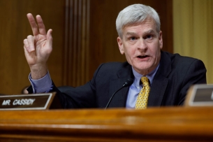 Louisiana senator Bill Cassidy, a white man with gray hair, wearing a suit, blue shirt and yellow tie, raises two fingers during a Senate hearing. 