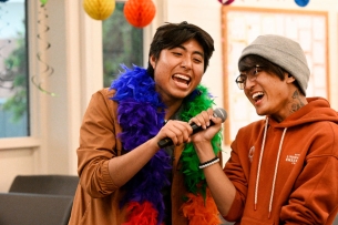 Two students sing karaoke surrounded by rainbow decorations, one of them wearing a rainbow boa
