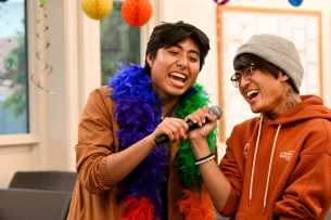 Two students sing karaoke surrounded by rainbow decorations, one of them wearing a rainbow boa