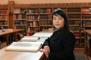 A photo of Bandy X. Lee, a light-skinned woman with dark hair and bangs, leaning on a shelf in a library. 