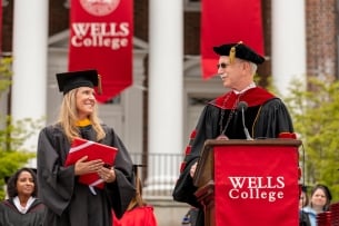 Wells College president Jonathan Gibralter, a light-skinned man with gray hair and a mustache, wearing academic regalia and sunglasses, on graduation day in front of red Wells College signage.