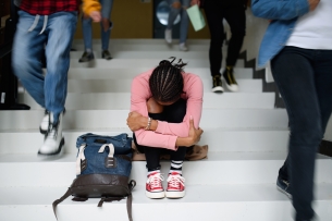A student with her dark hair in braids sits on a staircase, hugging her arms around herself, as other students walk past