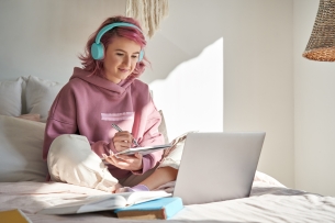 A female student sits on her bed wearing headphones taking notes during an online class.