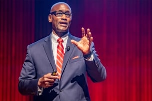 A dark-skinned man in a suit and tie and glasses speaks in front of a red curtain