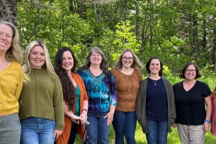 College and career success coordinators from Maine community colleges stand together smiling at a summer retreat with trees in the background.