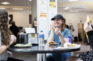 Three female students sit at a small table in a cafeteria eating sandwiches, laughing. 