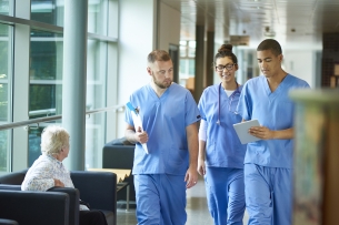 Three students in blue scrubs walk down a hallway in a hospital