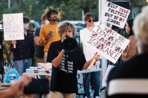 A young white man with dark blond hair holds a megaphone and a sign that says "I won't be censored to comfort your ignorance."