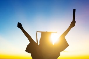 A graduate with a diploma sticks their arms out in celebration facing the sunset.