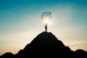 Silhouette of person standing on a mountain and holding a target with an arrow in the bull's-eye, with the sun going down behind them 