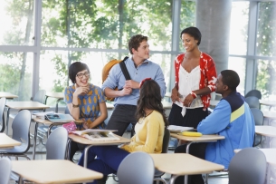 A group of diverse college students chat sitting at desks in a college classroom.