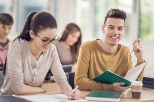 Two students sitting next to each other in class are writing. Their phones are not visible.