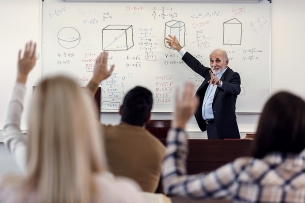 Math instructor at the whiteboard with students in front of him raising their hands.