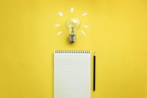 Pad of paper and pencil on  a yellow background with a bright light bulb above them