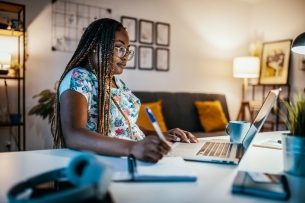 An African American female student takes notes in front of a laptop, engaging in online learning.