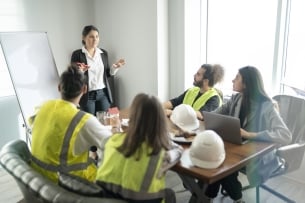 A woman in a suit jacket with a whiteboard stands in front of a group of four students wearing yellow vests and construction hats. 