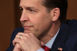 A close-up of Ben Sasse's face, resting his chin on this folded hands. He is a light-skinned man with short dark hair who is wearing a suit.