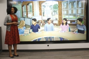 A woman stands next to a screen with images of children sitting at a table in a classroom setting 
