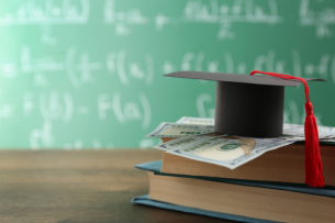 A stack of books, topped by cash and a graduation cap, in front of a chalkboard.