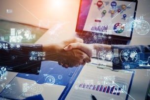 Two male hands shake while there are blue "digital" icons floating around. A computer screen and paper bar chart are in the background.