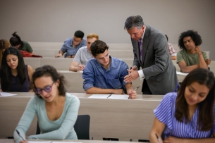 A professor speaks to a student in his lecture hall class, as other students are working on assignments.